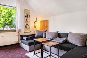 A modern living room with a sofa, two side tables, a decorative wall piece, and a window with a view of greenery. - Holiday Apartment 'Zum Gingko' with Balcony (Neuenkirchen)
