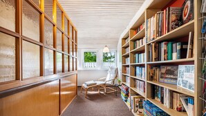 A library with bookshelves, a reading chair, and a window with a view of trees.
