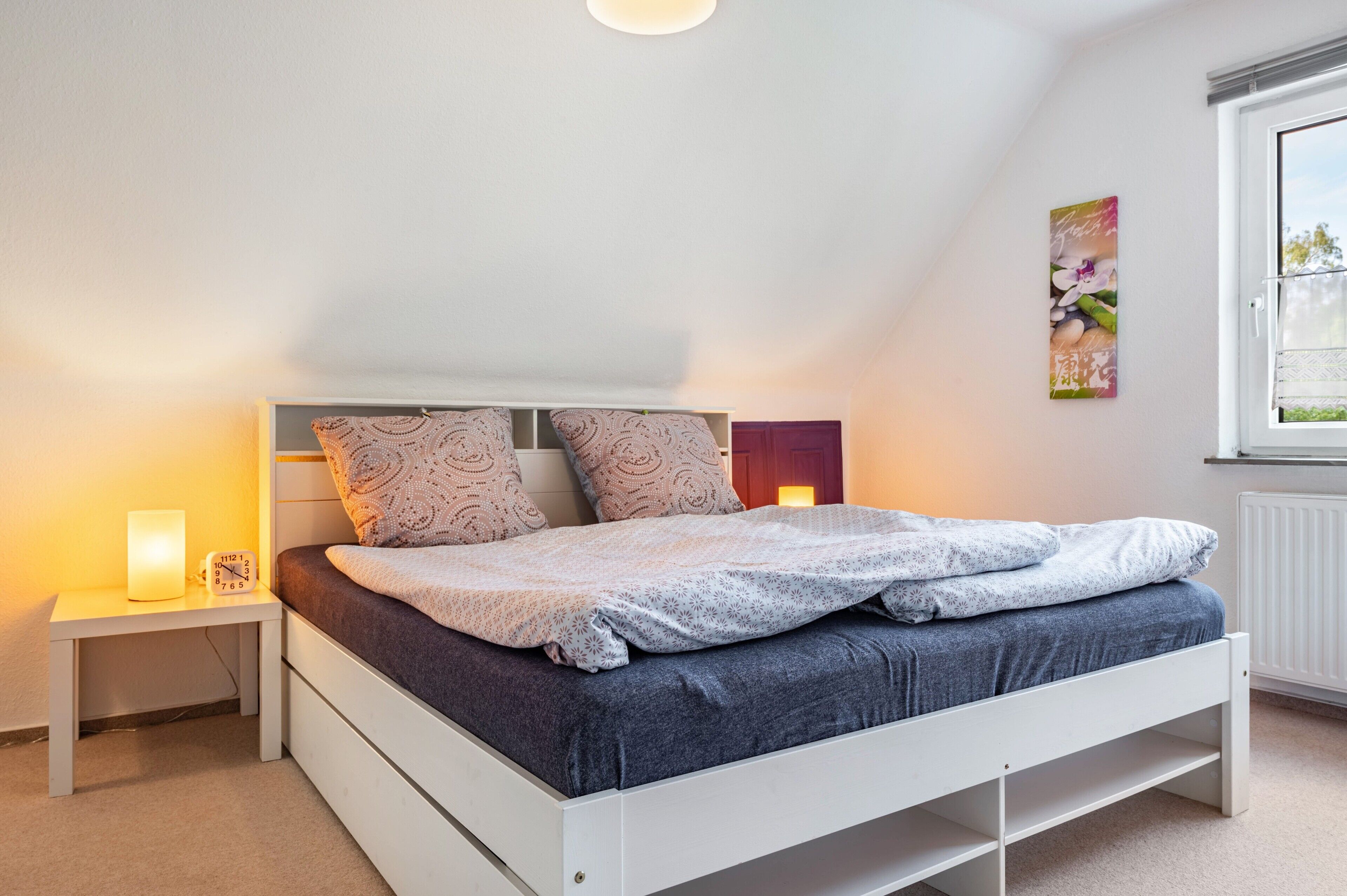 Un dormitorio con cama, mesita de noche, una obra de arte en la pared y una ventana.