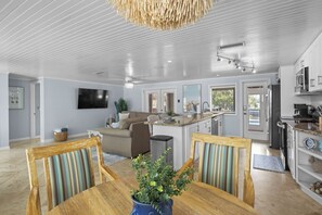 Eine moderne Küche mit weißen Schränken, ein Essbereich mit gestreiften Stühlen sowie ein Wohnzimmer mit Sofa und Fernseher.