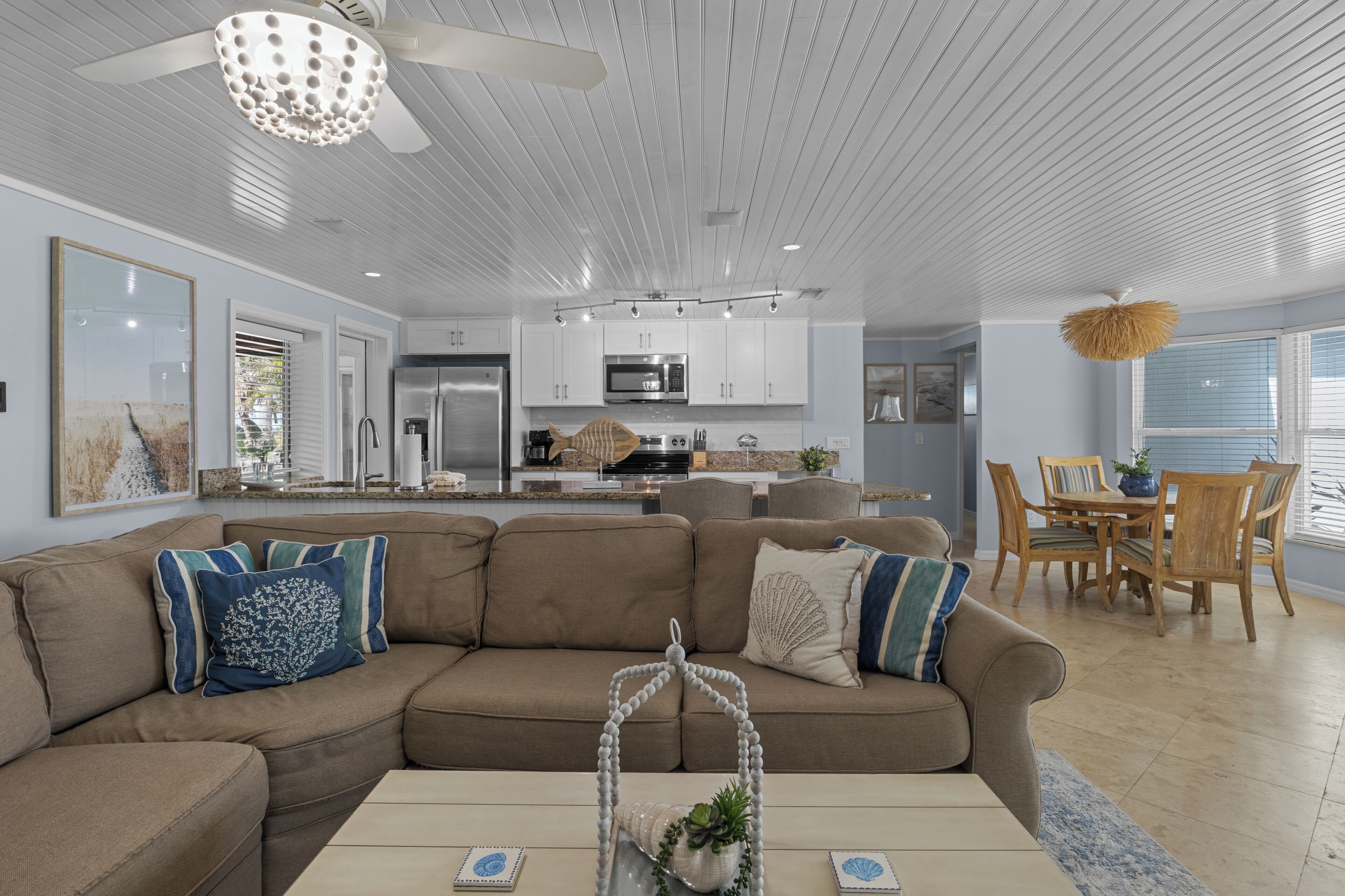 A modern living room with a sectional sofa, a dining area with wooden chairs, a kitchen with white cabinets, and a ceiling fan.