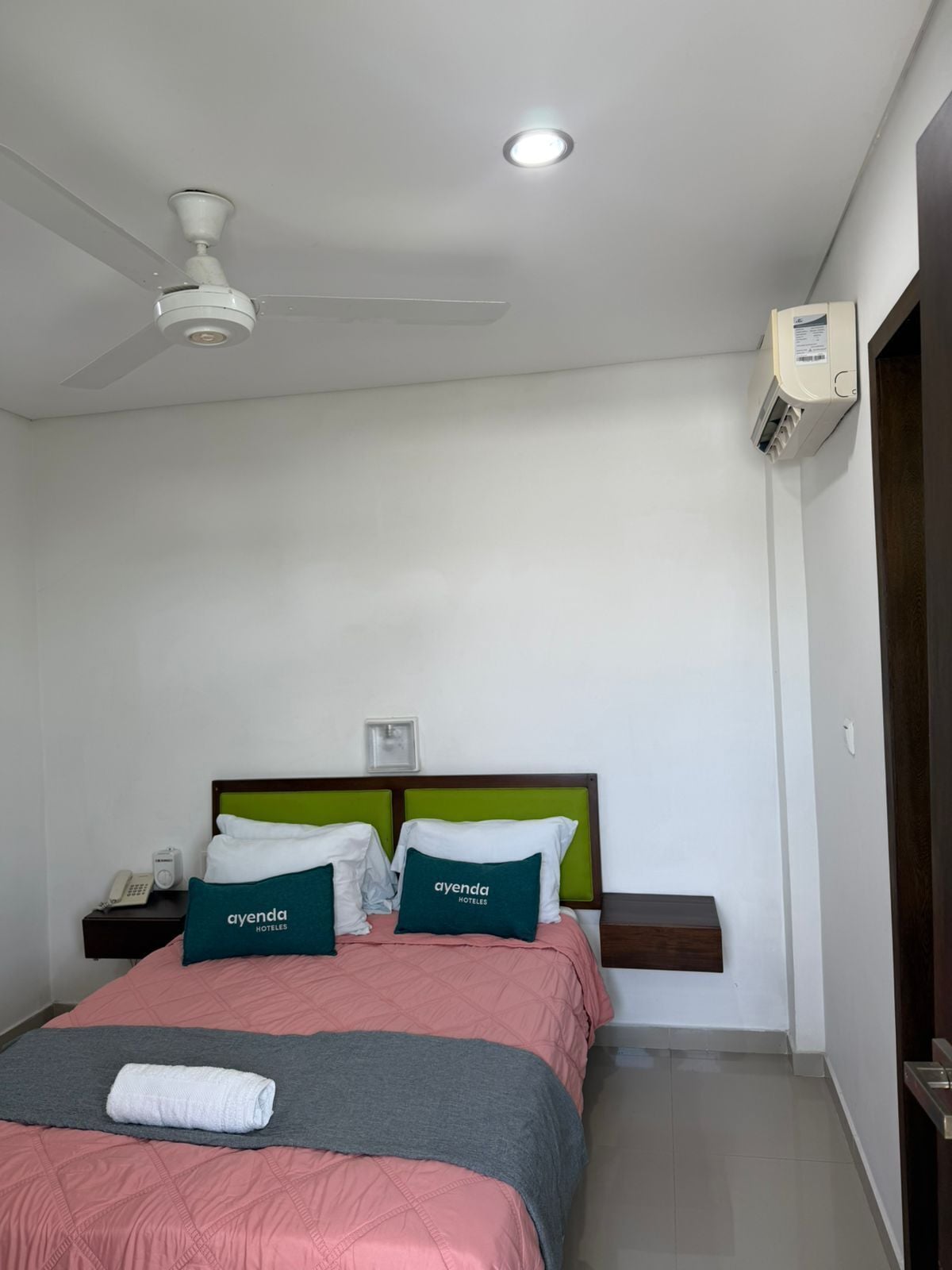 Una habitación de hotel con cama, mesita de noche, teléfono, ventilador de techo y unidad de aire acondicionado.