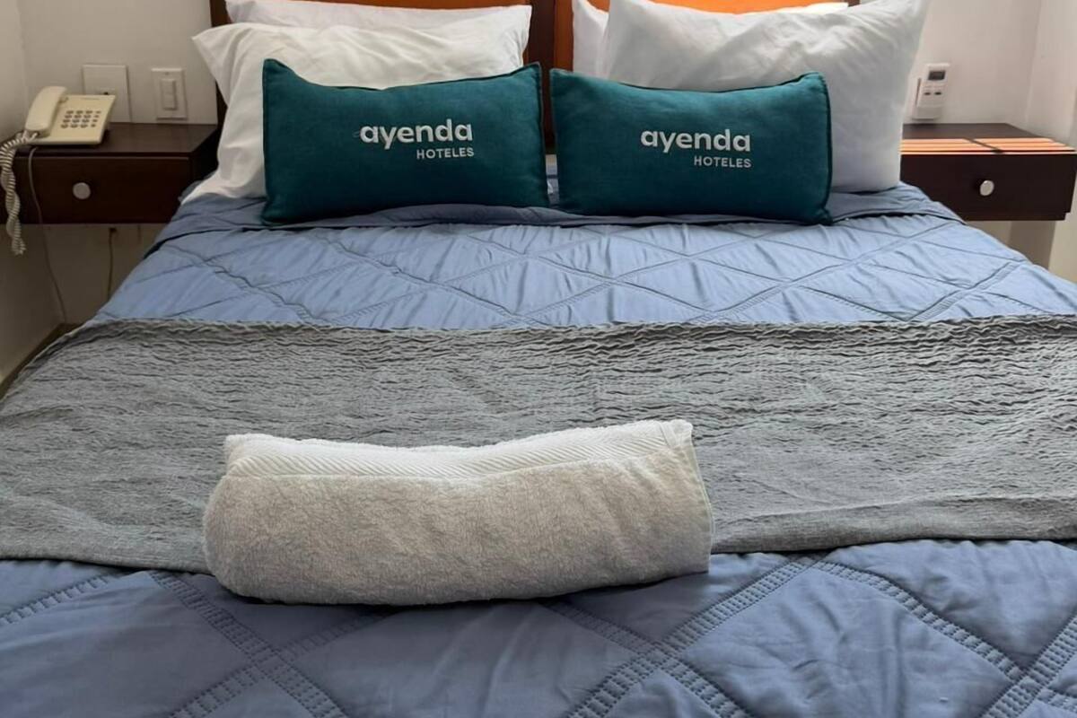 Una habitación de hotel con cama, dos almohadas con la marca 'ayenda HOTELES', teléfono en la mesita de noche y ventana con vista al cielo.
