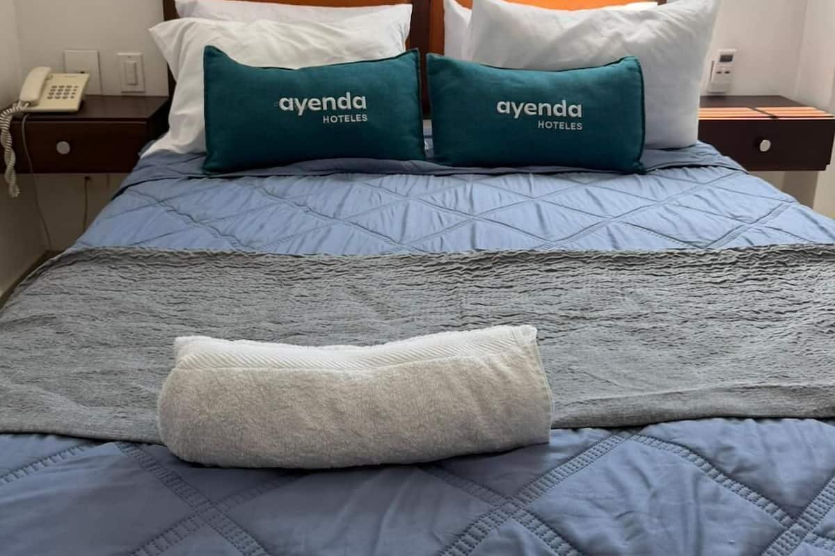 Une chambre dâhĂŽtel avec un lit, deux oreillers portant la marque « ayenda HOTELES », un tĂ©lĂ©phone sur la table de chevet et une fenĂȘtre donnant sur le ciel.