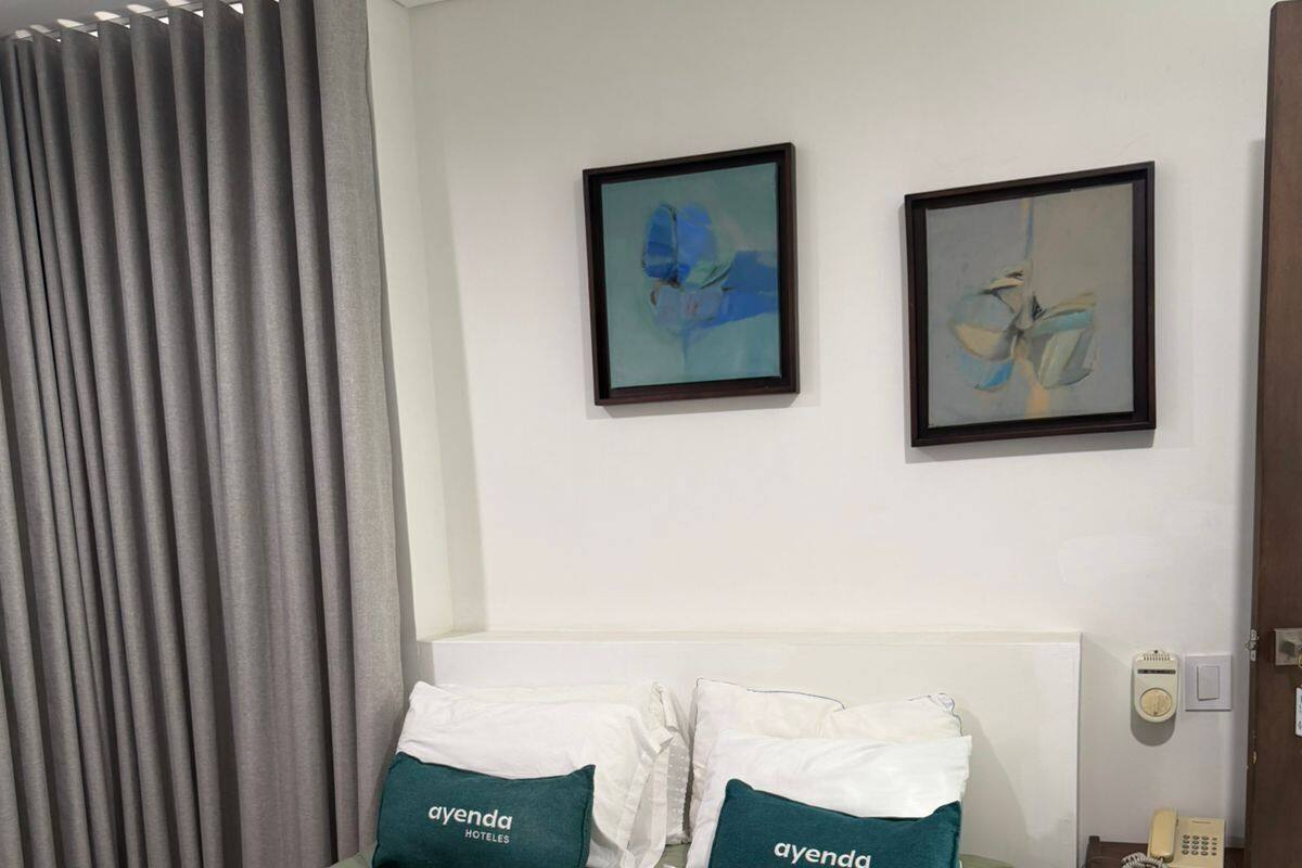 Una cama bien hecha con una colcha verde, dos almohadas con la marca 'ayenda hotels', una manta blanca y un teléfono sobre una mesita de noche.