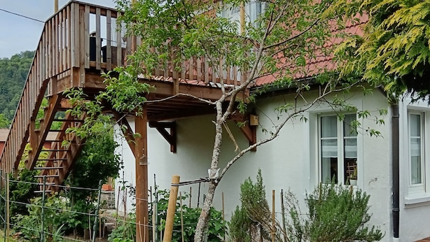 A house with a wooden staircase leading to a deck, surrounded by a garden with various plants and a fenced area. - In a house with garden, apartment with terrace near a lake. (Oderen)