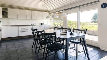 A modern kitchen with white cabinets, a dining table with chairs, and large windows offering a view of greenery.