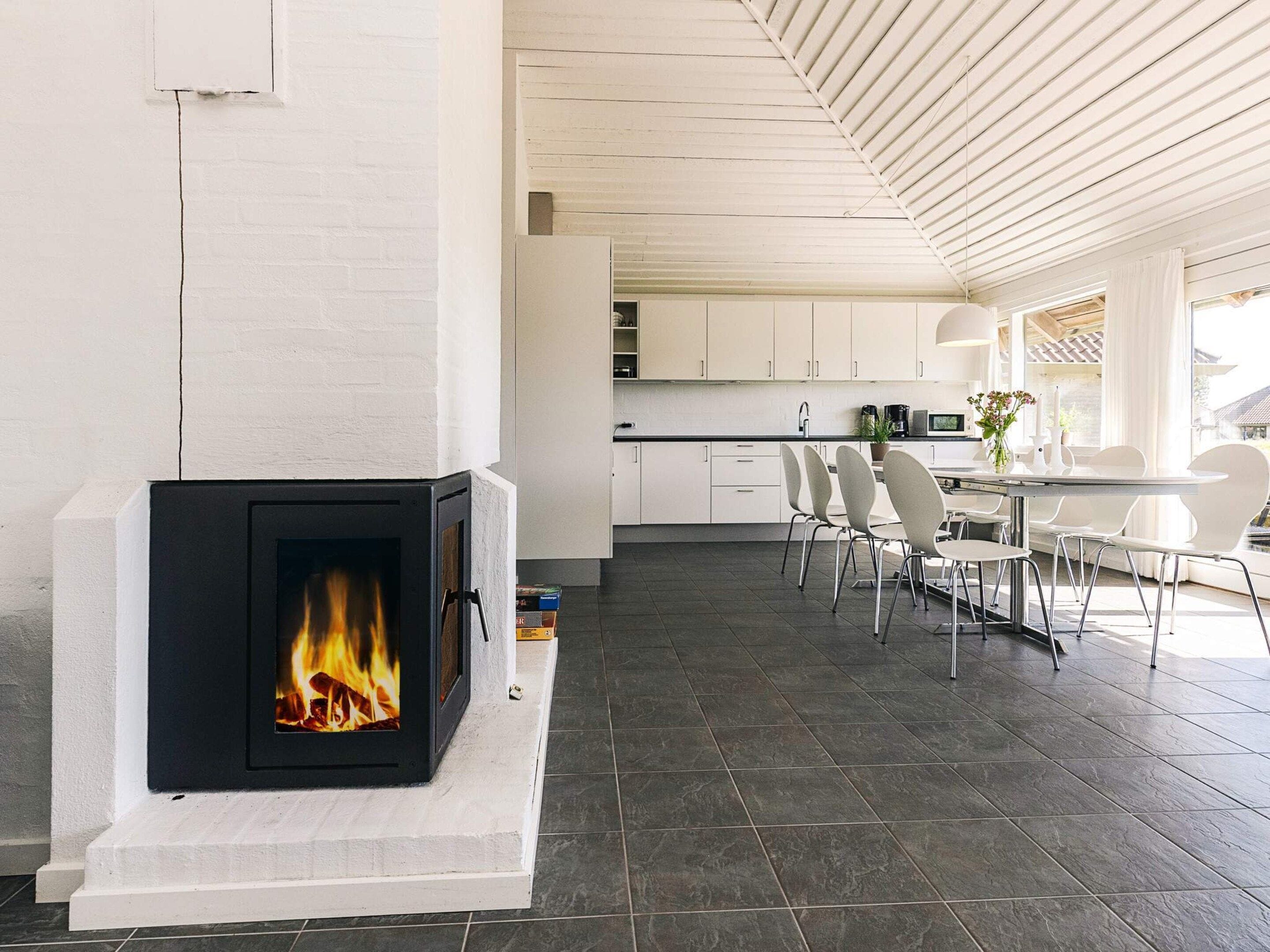 Une cuisine moderne avec des meubles blancs, une cheminée où l’on peut voir les flammes, et un coin repas avec des chaises et une table blanches.