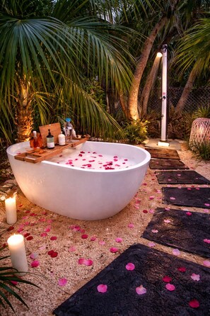 A large outdoor bathtub filled with water and rose petals, surrounded by a garden with palm trees and a pathway.