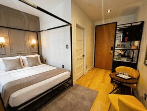 Ein Schlafzimmer mit Himmelbett, einer kleinen Essecke mit einem gelben Stuhl und einer Küchenzeile mit einem schwarzen Regal.
