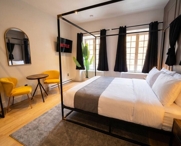 Ein modernes Hotelzimmer mit einem Himmelbett, einem runden Tisch, zwei gelben Stühlen, einem großen Spiegel und einem Fernseher mit Netflix.