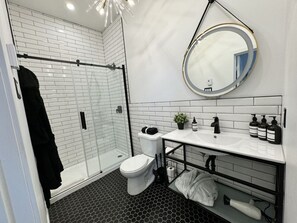 Ein modernes Badezimmer mit Duschkabine aus Glas, einer weißen Toilette, einem schwarz-weißen Fliesenboden und einem runden Spiegel über dem Waschbecken.