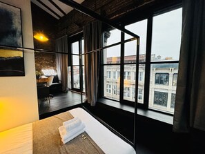 Ein modernes Hotelzimmer mit großem Fenster, einem Bett, einem kleinen Tisch und Blick auf die Stadtlandschaft.