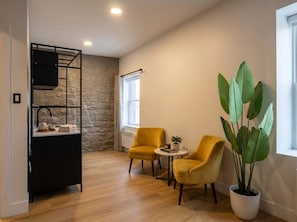 A room with a stone wall, a black metal shelf, two yellow chairs, a small table, and a large potted plant. - Boutique Old Montreal Suite-1C (Montreal)