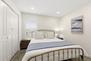 A bedroom with a bed, a nightstand, a dresser, a window with blinds, and a painting on the wall. - Claiborne Inn (Lafayette)