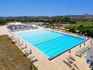 Una gran piscina rectangular con agua cristalina azul, rodeada de sillas de descanso y sombrillas.