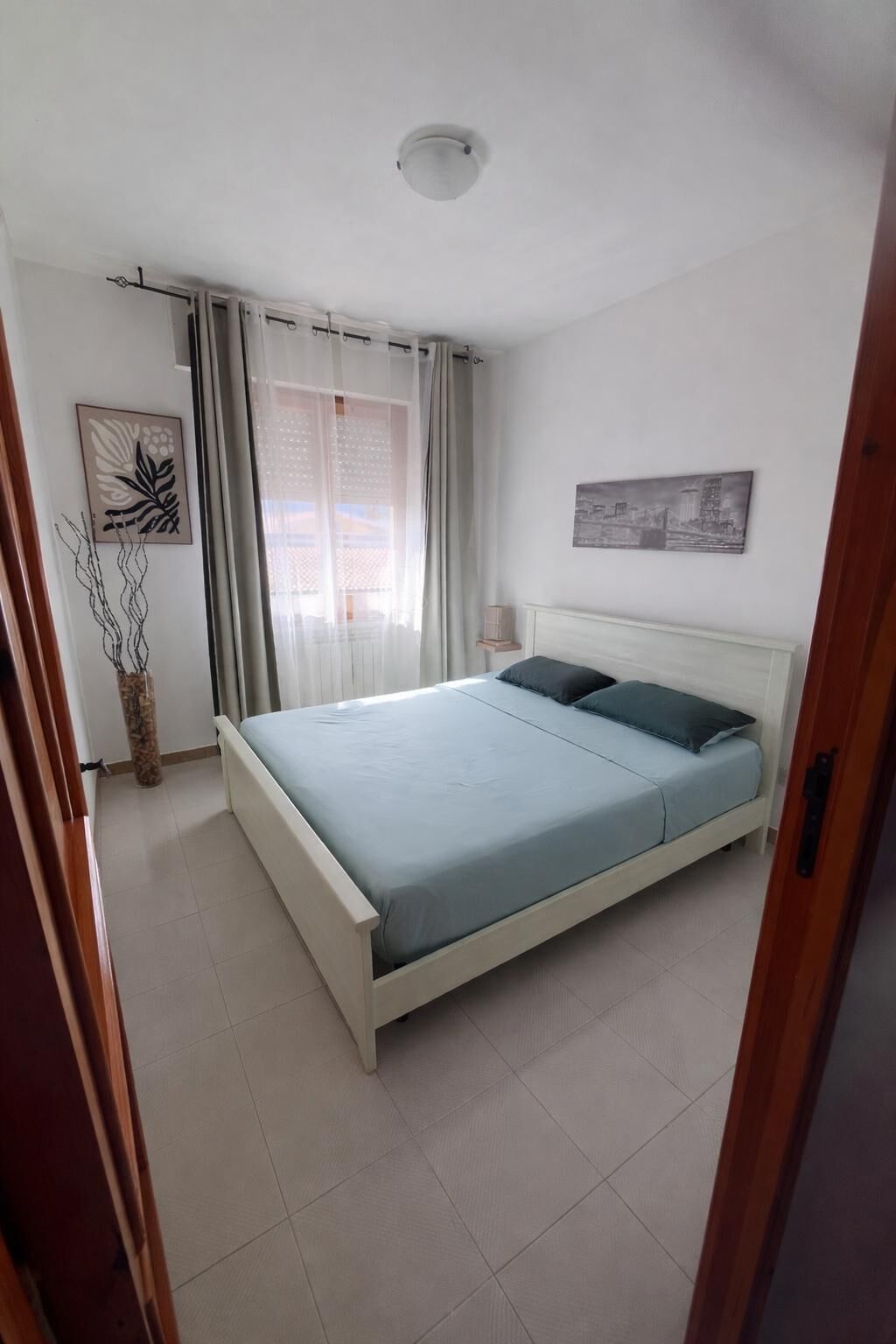 Un dormitori amb llit, cortines, làmpada penjada a la paret i una pintura de paisatge urbà.