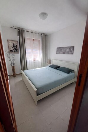 A bedroom with a bed, curtains, a wall-mounted lamp, and a cityscape painting. - Valledoria House - Two Bedroom Apartment, Sleeps 4 (Valledoria)