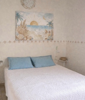 A bedroom with a bed, two pillows, a wall-mounted painting of a beach scene, and a shelf with a basket. - Villa Aura - Two Bedroom Apartment, Sleeps 6 (Pirotto Li Frati)