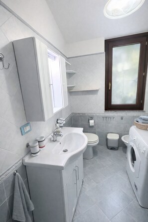 Ein modernes Badezimmer mit Waschbecken, zwei Toiletten, einer Waschmaschine und einem Fenster.