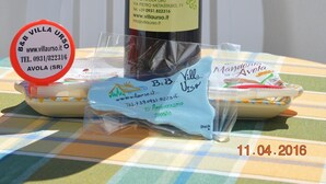 A bottle of wine, a small plate with food, and a plastic bag with a label on a table with a view of the sea. - B&B Mare e Sole Villa Urso (Avola)