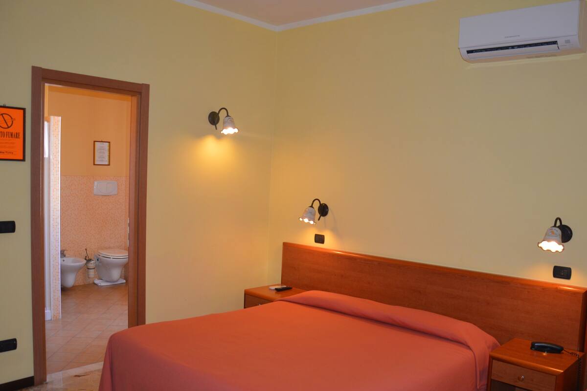A hotel room with a bed, bedside tables, an air conditioning unit, and a bathroom visible through an open door.