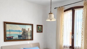 Un dormitorio con cama, mesita de noche, un cuadro, ventana con cortinas y lámpara colgante.