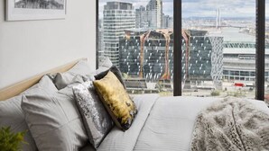 Un dormitorio con cama, almohadas, una mesita de noche con vela y un ventanal con vistas a la ciudad.