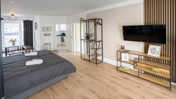 Et moderne hotelværelse med en stor seng, et fjernsyn, et træ-tv-bord, et skrivebord og et køkkenområde i baggrunden.