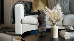 Un salon moderne avec une table basse ronde noire, un vase blanc contenant des fleurs séchées, un canapé gris et blanc agrémenté d’un plaid, et un grand lampadaire doté de trois abat-jour sphériques.