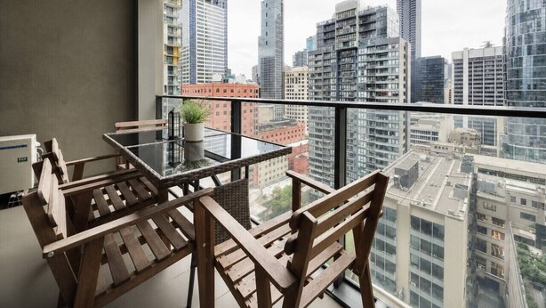 Un balcon avec une table en verre et des chaises en bois, donnant sur un paysage urbain avec de hauts immeubles.