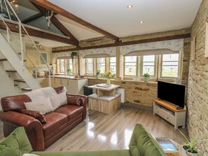 A modern living room with a brown leather sofa, a green armchair, a wooden coffee table, a TV stand, and a staircase leading to an upper level. - Riverview Cottage (Holmfirth)