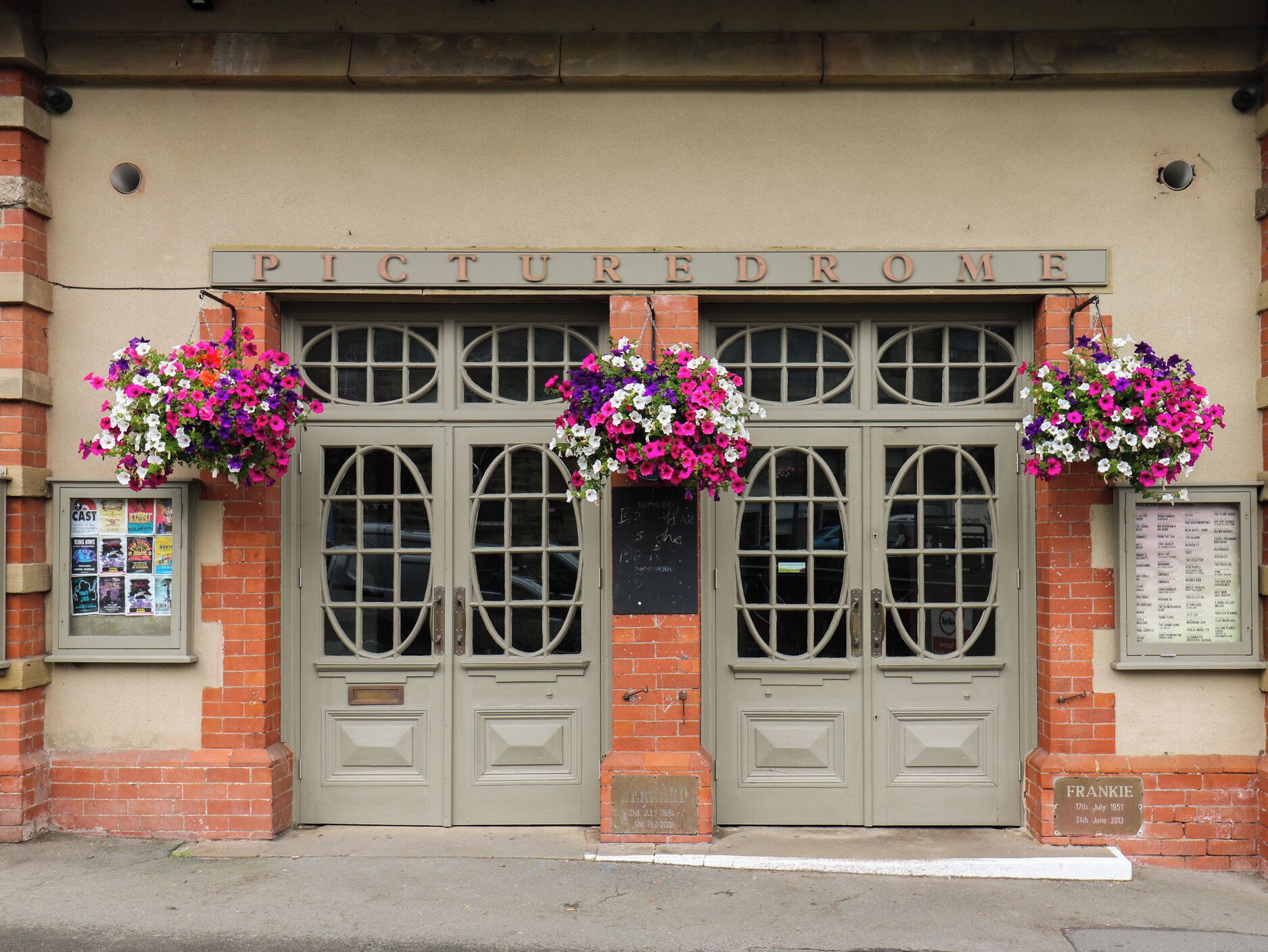 Eine Gebäudefassade mit einem Schild, auf dem 'PICTUREDROME' steht, mit Doppeltüren und runden Fenstern sowie hängenden Blumenkörben.