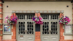 Eine Gebäudefassade mit einem Schild, auf dem 'PICTUREDROME' steht, mit Doppeltüren und runden Fenstern sowie hängenden Blumenkörben.