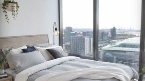 A modern bedroom with a large window, a bed with grey bedding, a bedside lamp, and a hanging plant. - Urban Skyline Escape (Melbourne)