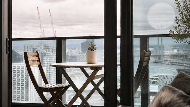 A balcony with a view of a cityscape, a chair, a small table with a plant, and a large window. - Urban Skyline Escape (Melbourne)
