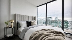 A modern bedroom with a large bed, a side table, a lamp, and a view of a cityscape through large windows. - Central Melbourne Skyline Retreat (Melbourne)