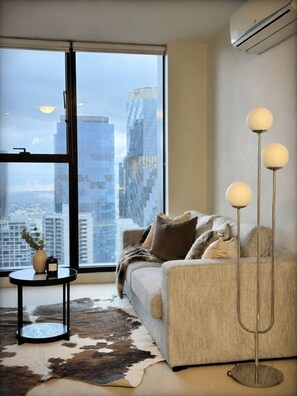 A modern living room with a sofa, a round coffee table, a floor lamp, and a city view through large windows. - Skyline Luxe in Melbourne CBD (Melbourne)