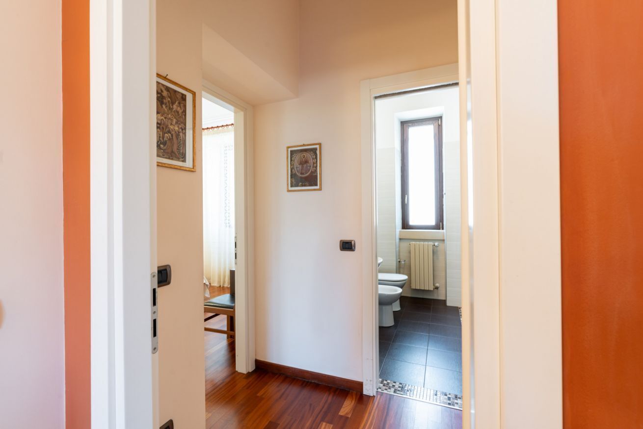 Un corridoio con pavimento in legno, una porta che dà su un bagno con water e termosifone, e un quadro appeso al muro.