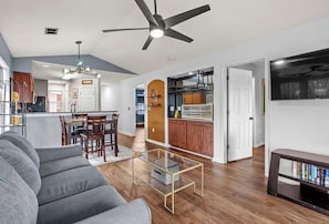 Ein modernes Wohnzimmer mit Sofa, Couchtisch und einem TV-Schrank. Im Hintergrund ist ein Essbereich mit Stühlen und eine Küche erkennbar.