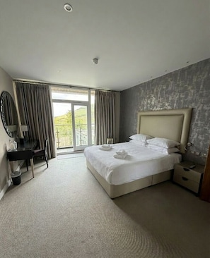 A hotel room with a large bed, a desk, a chair, a mirror, and a view of the outdoors.