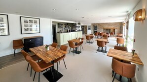 A modern hotel dining area with multiple wooden tables and chairs, a kitchen in the background, and framed artwork on the walls. - Llawnroc Hotel (Gorran Haven)