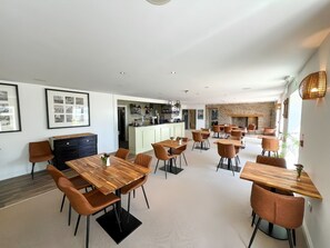 A modern hotel dining area with multiple wooden tables and chairs, a kitchen in the background, and framed artwork on the walls.