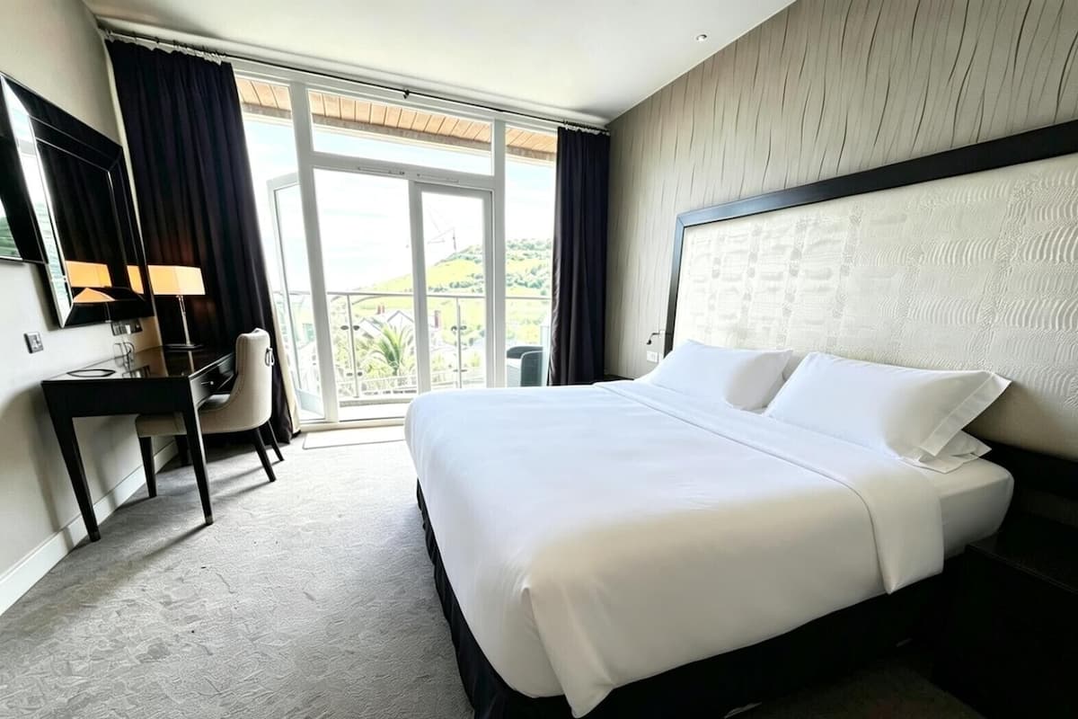 A hotel room with a large bed, a desk, a chair, and a balcony with a view.