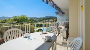 A balcony with a table set for two, chairs, a pool in the background, and a view of the countryside. - Roseraie Apartment with Private Pool, Wi-Fi, and Air Conditioning (Antibes)