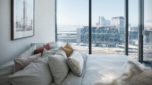 Een slaapkamer met een groot bed, meerdere kussens, een lijst met een afbeelding aan de muur en uitzicht op de stad door grote ramen.