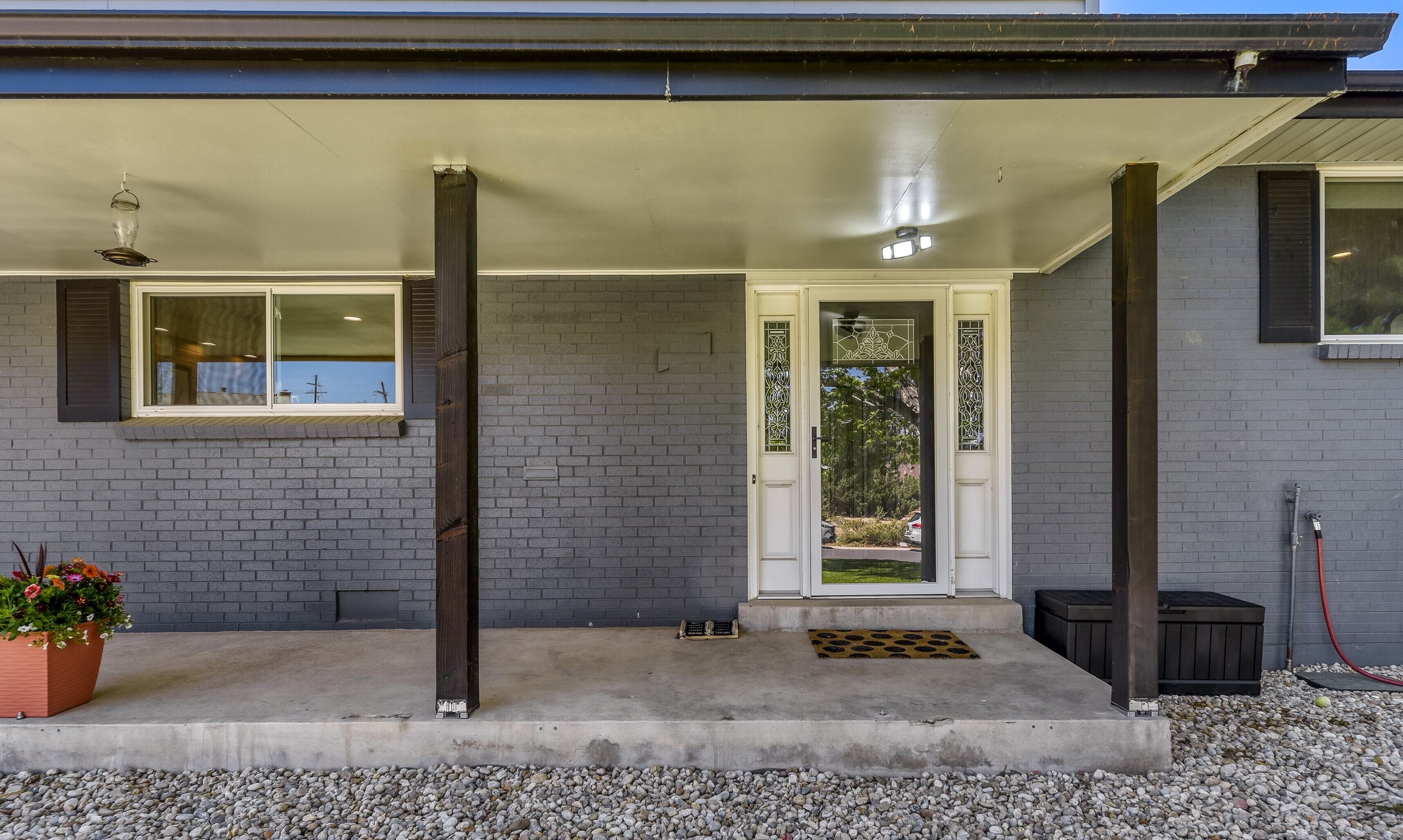Une maison avec un porche en béton, un mur en briques, une porte blanche ornée d’un panneau de verre décoratif et une fenêtre avec une grille en métal.