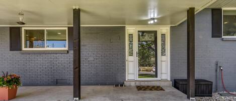Une maison avec un porche en béton, un mur en briques, une porte blanche ornée d’un panneau de verre décoratif et une fenêtre avec une grille en métal.