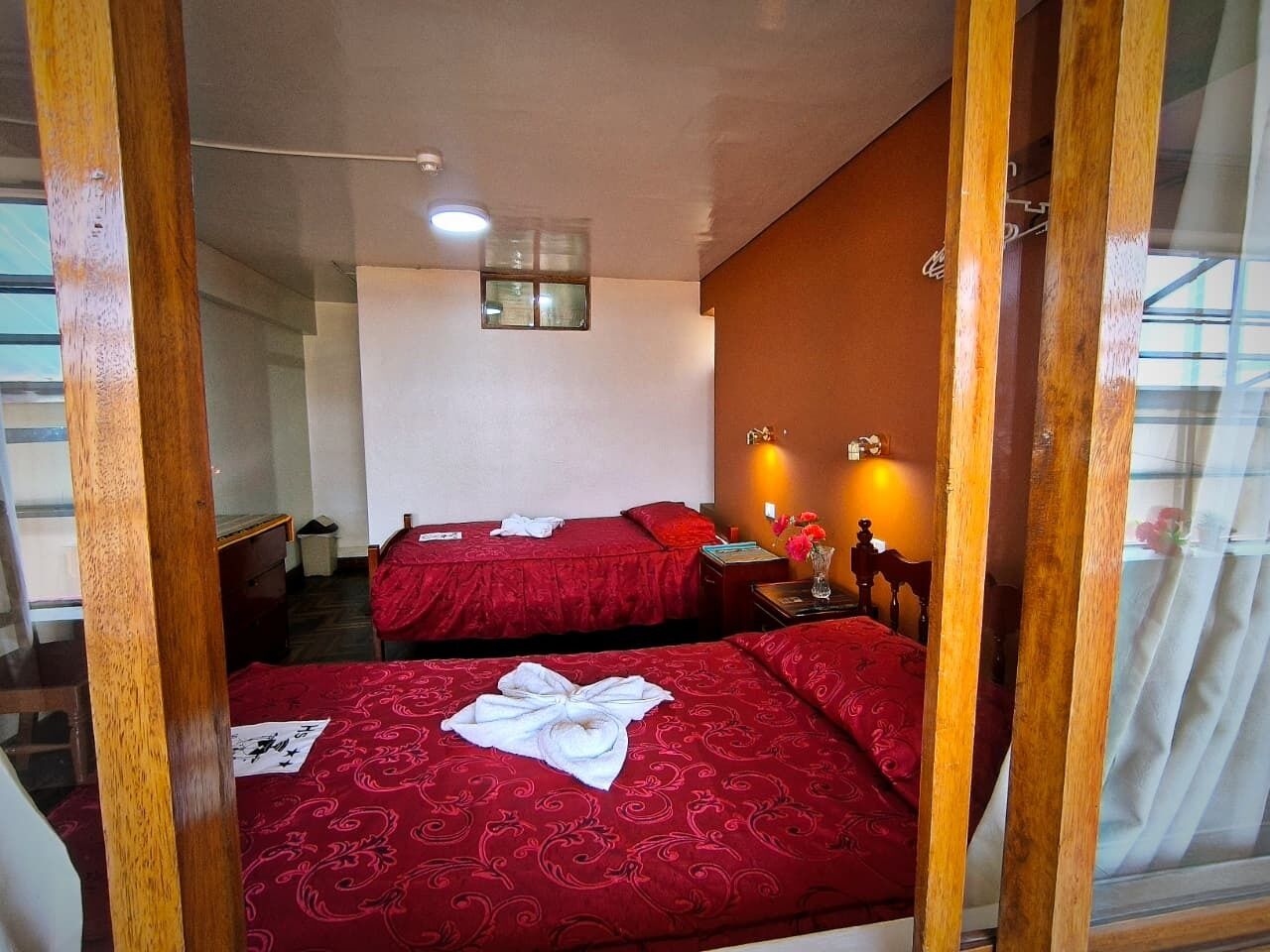 A hotel room with two beds, a small table, a nightstand, a window, and a wall-mounted light fixture.