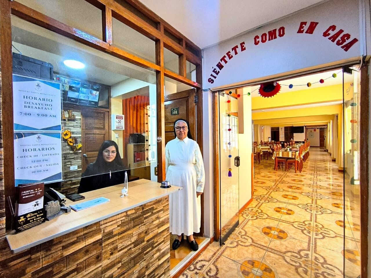 Un espace d’accueil d’hôtel comprenant un comptoir d’enregistrement, un réceptionniste et une salle à manger visible à travers l’entrée.